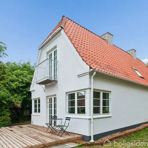 Hvidt hus med rødt tegltag, står stille på en træterrasse. To stole og et bord i forgrunden. Grønt buskads omkranser baggrunden under en blå himmel.