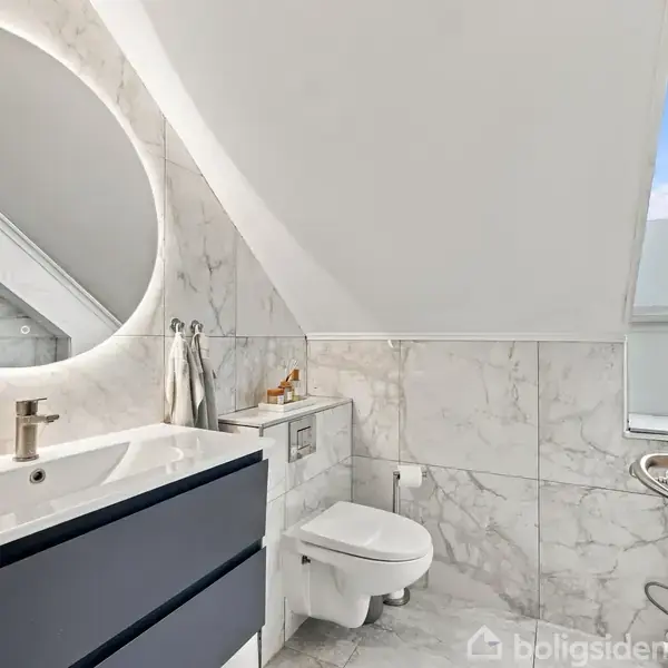 Badeværelse med vask og rund spejl på væggen, væghængt toilet ved siden af under et skråt loft i et rum med marmorerede vægfliser og et vindue til højre.
