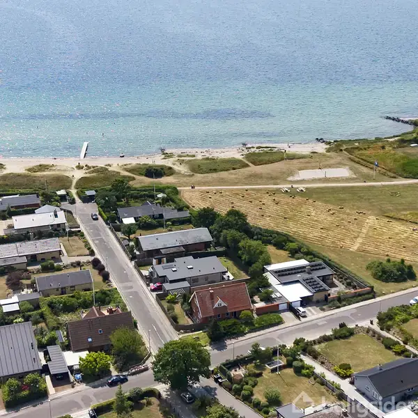 Boligområde med huse og haver nær stranden. Vejen fører til en kystlinje med en mole. Havet ligger roligt i baggrunden, omkranset af græsarealer og bebyggelse.
