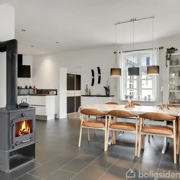 En brændeovn brænder i et moderne, lyst køkken-alrum med sorte flisegulve. Et langbord med stole står ved et vindue, omgivet af lyse møbler og væghængte lamper.