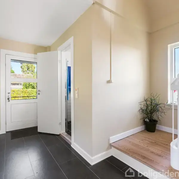 Dør står åben i en lys entré med sorte gulvfliser. Til højre ses en indvendig trappe, en tør plante i en krukke ved et stort vindue.