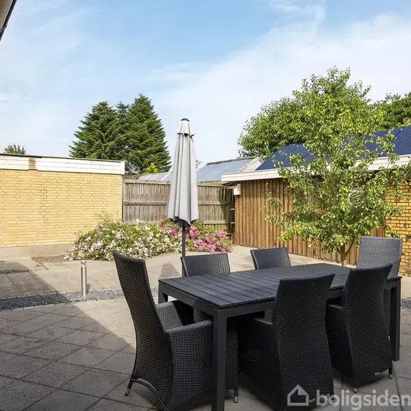 Et havebord med seks stole står på en flisebelagt terrasse ved siden af et sammenklappet parasol. Omkring er der en gul murstensbygning, blomstrende buske og en blå himmel.