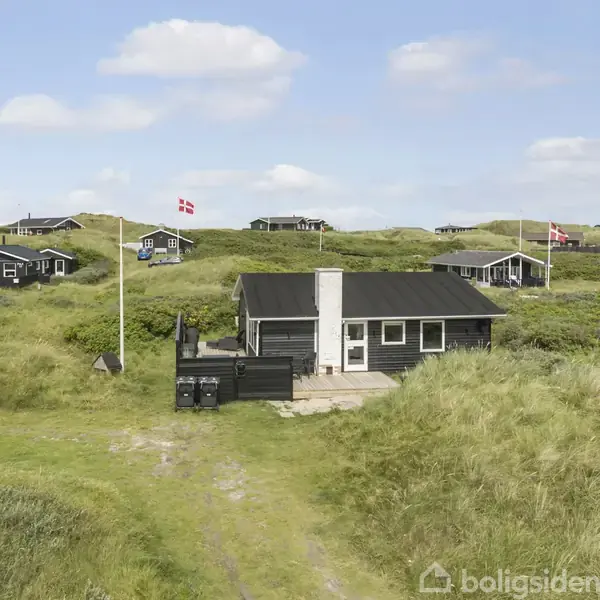 Et sort træhus med hvidt tag står stille blandt græsbeklædte klitter. I baggrunden ses flere sommerhuse og flag, der markerer en dansk kystoplevelse.