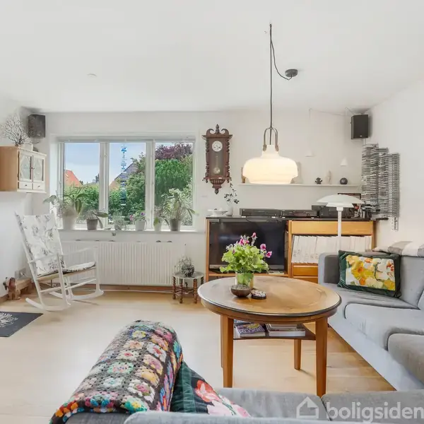 Stue med grå sofaer og rundt træbord med blomster. Uret hænger på væggen. Vindue med udsigt til have. Lys hænger fra loftet. Maleri hænger på væggen.