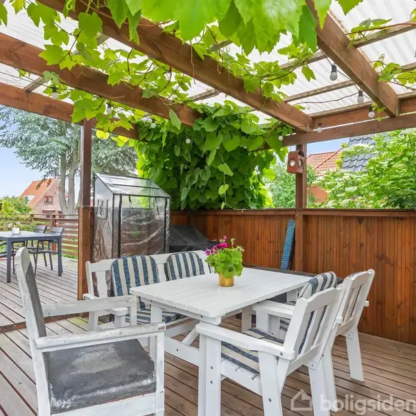 Hvidt bord med stole står under en pergola med grønne blade. Omkring er der træhegn og terrasseplanker. En potteplante med blomster pynter på bordet, og det er solrigt udenfor.