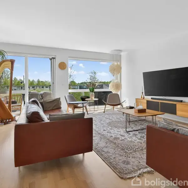 En elegant stue med en brun lædersofa og et sofabord centreret på et orientalsk tæppe. En harpe står ved vinduet, som åbner til en balkon med udsigt over grønne omgivelser. En fladskærm er monteret på en hvid væg over et træmøbel.