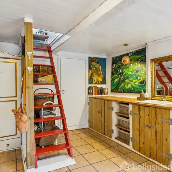 Et rum med en rød trappe, der fører opad. Til venstre ses et hvidt skab, og til højre er der et træbord med kunst på væggene.