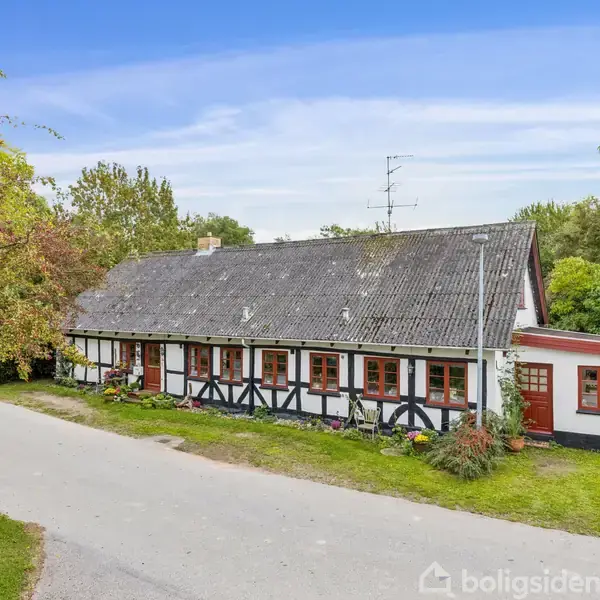 Bindingsværkshus med røde vinduer og sort tag står ved en stille landevej. Omringet af grønne træer og buske. Golvet er græs og der er en blå himmel.