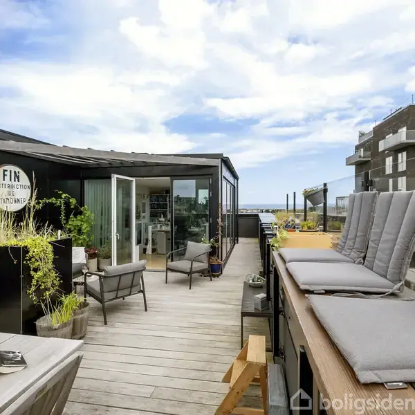 En moderne tagterrasse med liggestole og planter placeret omkring. Døre til en glasindgang er åbne. En rund sign indeholder teksten: "FIN i det fri stations". Bygninger ses i baggrunden.