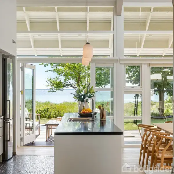Køkkenø med frugtskål står i et moderne, lyst køkken med hvide vægge og store vinduer. Døre åbner ud til terrasse med udsigt over en frodig have og havet i baggrunden.