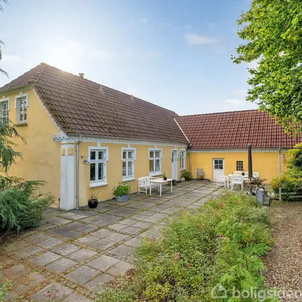 Gul bygning med rødt tag ligger i solbeskinnet gårdhave med fliser og havemøbler, omgivet af grønne buske og træer. Ingen synlig tekst.