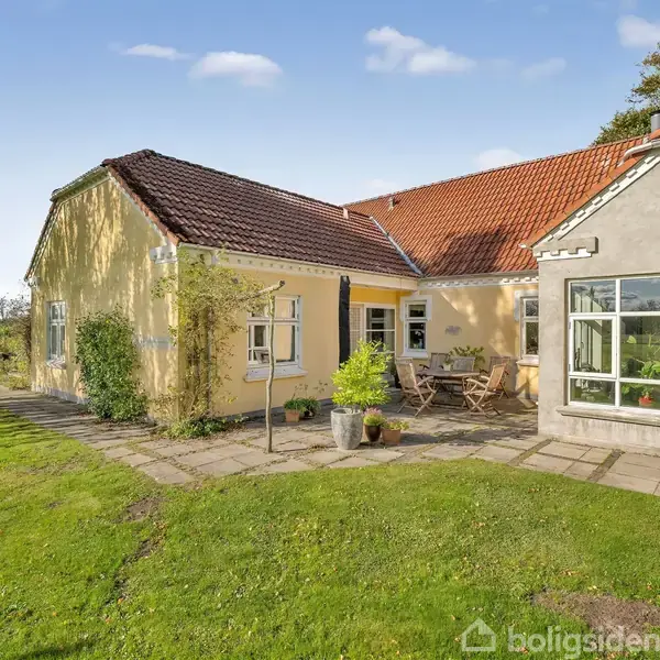 Et gult hus med rødt tegltag står i solskin. Foran huset er en terrasse med havemøbler. Græsplæne og træer omgiver huset i en rolig landskabelig have.