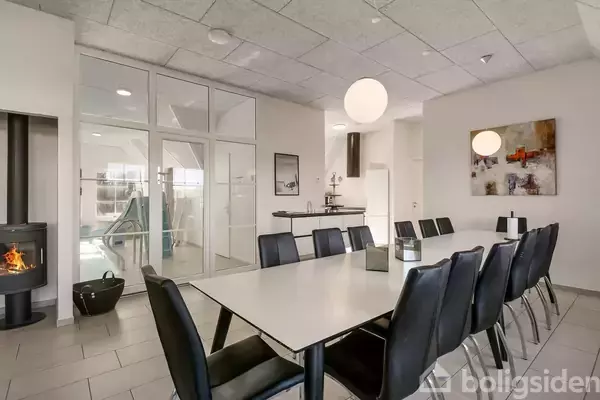 Langt bord med sorte stole står i et lyst rum. Pejs til venstre brænder, og store glasdøre leder til en anden del af huset. Kunst hænger på væggen.