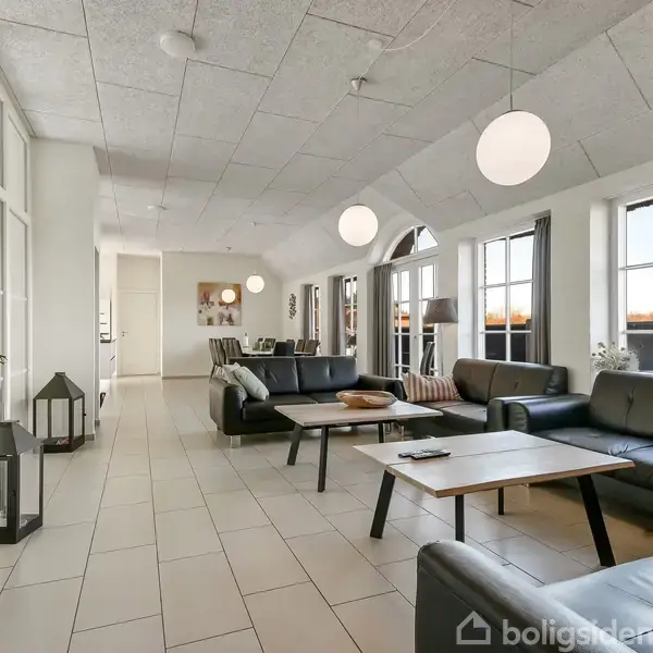 Stue med sorte lædersofaer omkring et lavt træbord. Store vinduer giver lysindfald, mens hængende kuglelamper supplerer belysningen. Dansk flag på hylde, mørke gardiner ved siden af.