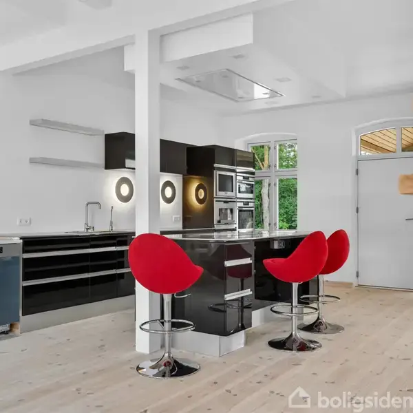 Køkken med sort højglansbord og røde barstole i moderne design. Køkkenø med indbygget ovn og mikrobølgeovn. Rummelig med lys træfarvet gulv og trappe til højre.