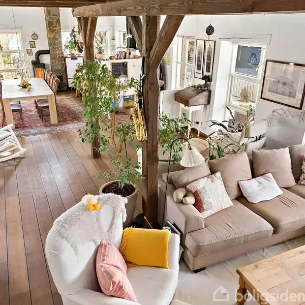 En stue med en stor hjørnesofa fyldt med puder ligger ved en rustik træsøjle. I baggrunden står et spisebord, omgivet af grønne planter og dekorative elementer i rummet.