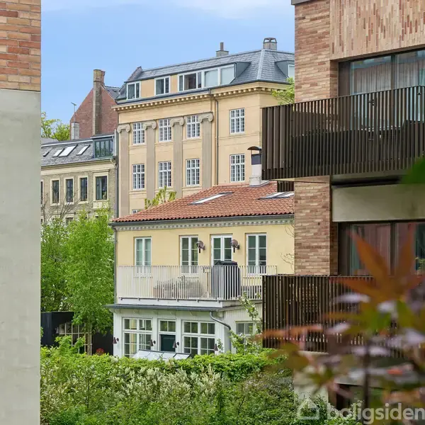 Beboelsesejendomme i moderne stil med store vinduer, omkranset af grønne buske og træer, placeret i en bynær beliggenhed. Himlen er blå med få skyer.