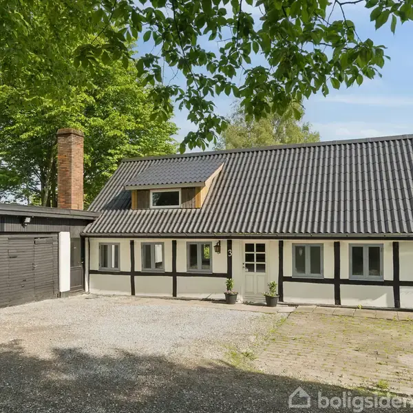 Et bindingsværkshus med sort tag og garage står på en gårdsplads med grus. Træer i baggrunden giver et grønt element til omgivelserne.