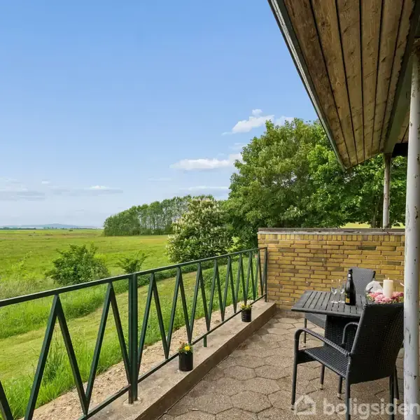 Veranda med sorte stole og bord står under et tag med udsigt over grønt landskab og skov i baggrunden. Sollys belyser området, der skaber en fredelig atmosfære.
