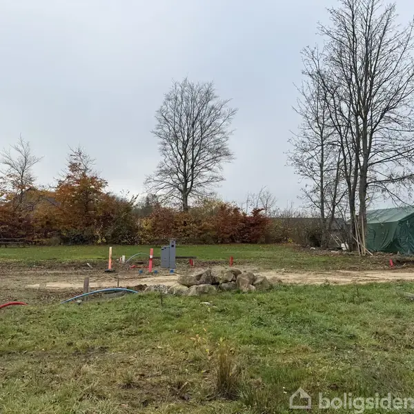 En byggegrund med jorden klar til konstruktion, omgivet af græs og træer. I baggrunden ses en grøn presenningsoverdække. Træerne er uden blade, og himlen er overskyet.