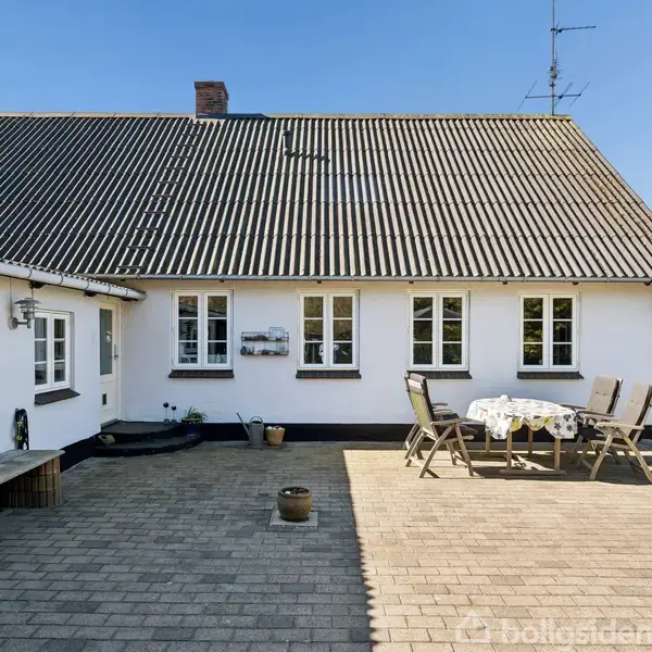 Huset har hvid facade og skråtag. En terrasse med bord og fire stole står i gårdhaven omgivet af fliser. Blå himmel og grønne træer kan ses i baggrunden.