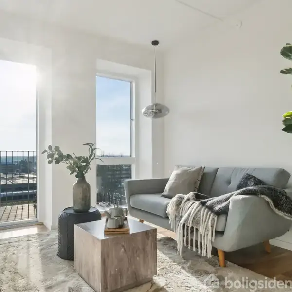 En stue med en grå sofa dækket af tæpper ved siden af et træbord med planter. Store vinduer slipper sollys ind, og en balkon er synlig udenfor.