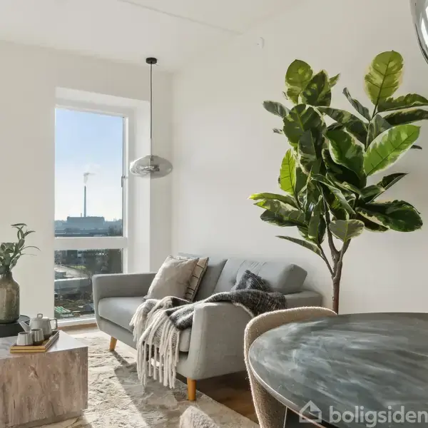 En lys stue har en grå sofa med tæpper, store grønne planter og et rundt bord i forgrunden. Store vinduer giver udsigt til en by og himmel i baggrunden.
