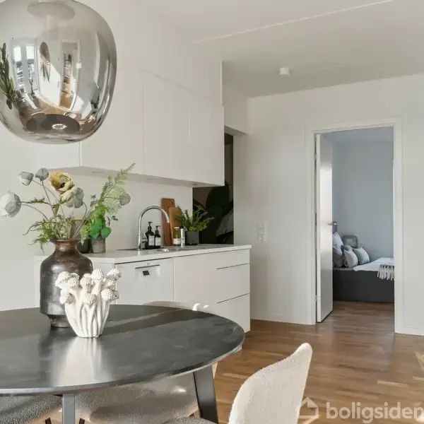 Rundt spisebord med blomsterdekoration, stående i et lyst køkkenområde. I baggrunden ses dør til soveværelse med seng, og der hænger en moderne lampe over bordet.
