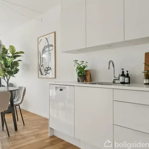 Hvidt køkken med vask og opbevaring, dekoreret med planter og sæbedispensere. Ved siden af står et spisebord med stole, omgivet af store planter og en moderne lampe.
