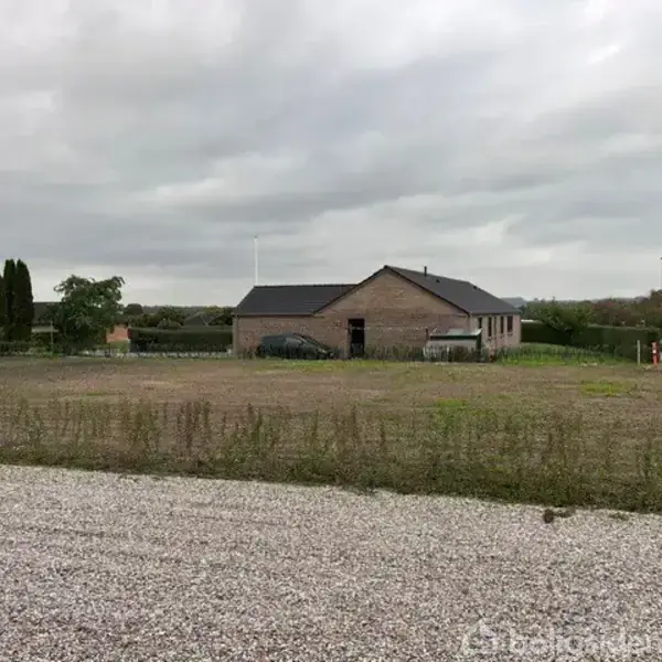 En murstensbygning står stille ved en grusvej, omringet af åbne græsarealer og nogle træer under en overskyet himmel. Der er et skilt ved vejen, men teksten er ikke læsbar.