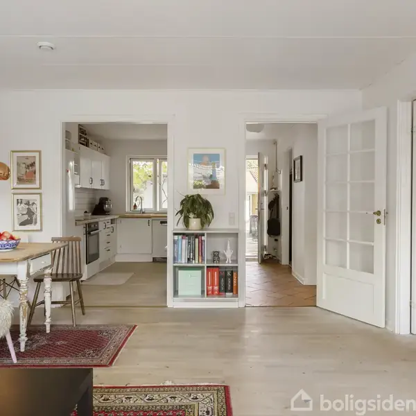Spisestue med rundt bord og stole pyntet med pels, placeret på tæpper. Køkken i baggrund med hvide skabe og stue til højre med reol. Moderne indretning.