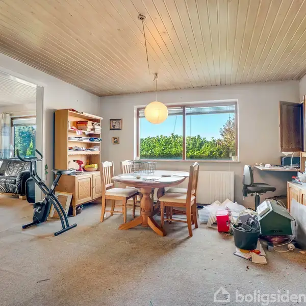 Et rodet rum med spisebord midt i, omgivet af stole. En motionscykel og bogreol står til venstre. Gardinerede vinduer lader dagslys ind, og papiraffald ligger spredt omkring.