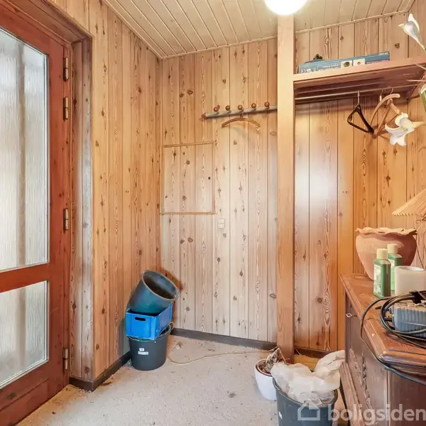 Dør til venstre åbner ind i en gang med træpaneler på væggene. Der står rengøringsartikler og en lampe på en trækommode. Flere kroge og hylder langs væggen.