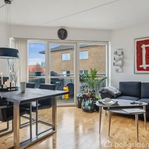 En stue med sort spisebord og stole ved siden af en mørk sofa. Vægkunst inkluderer et "Cirkel Kaffe" plakat og rød-hvidt japansk tegn. Store vinduer giver udsigt til en balkon.