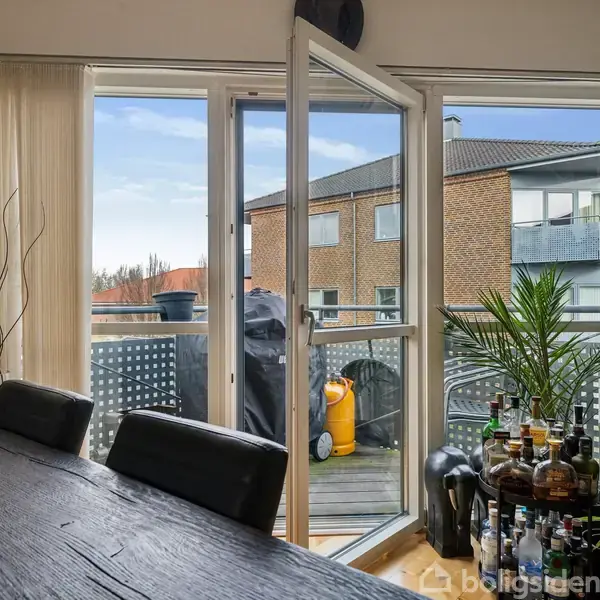 Et spisebord nær en åben altandør. Udenfor står en gasgrill og en gul gasflaske. Indendørs er der en plante og en samling af flasker.