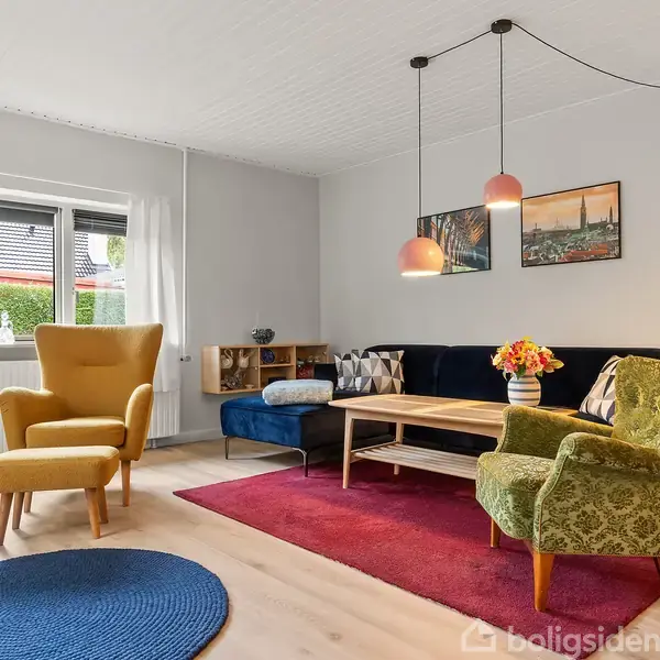 En stue med farverige møbler, inklusive en gul lænestol, grøn stol, og sort sofa, står på et trægulv med rødt og blåt tæppe, belyst af loftslamper.