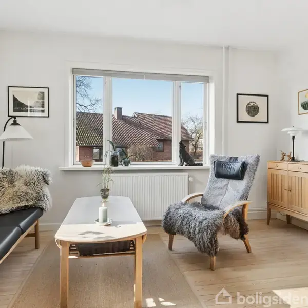 Stue med sort sofa og lænestol dækket af pelstæppe, står omkring et lavt sofabord. Væghængt ur og kommode i træ. Store vinduer viser tagene udenfor.