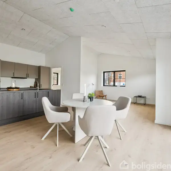 Et spisebord med fire hvide stole står i en lys, moderne stue. Til venstre er et åbent køkken med grå skabe. Store vinduer slipper lys ind.