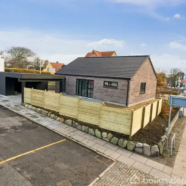 En moderne, murstensbelagt bygning med sort tag ligger ved en vej. Den er omkranset af et lavt træhegn og sten. Andre huse og en fortovsområde omkranser.