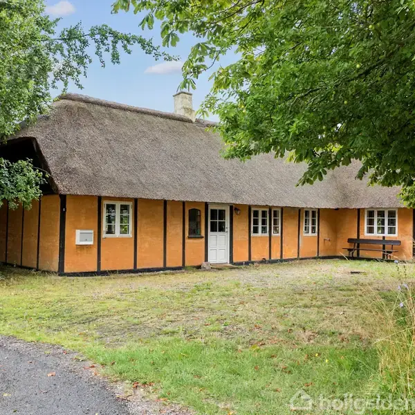 Stråtækt bindingsværkshus med orange facade og hvide vinduer står stille ved siden af en grusvej, omgivet af grønne træer og græsplæne. Bænkesæt foran huset.