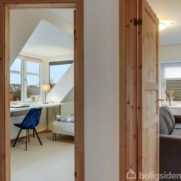 Et hyggeligt interiør med et skrivebord og en blå stol ved et vindue. Til højre ses en grå sofa i et tilstødende værelse. Begge rum har lyse trædøre.