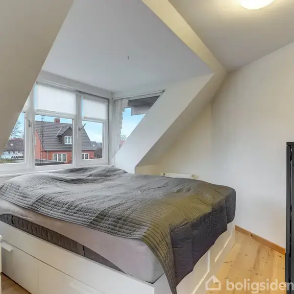 Seng placeret i et lyst værelse under en skråvæg, omgivet af en kommode med små planter og et stort vindue, der viser hustage udenfor. Ingen tekst til stede.