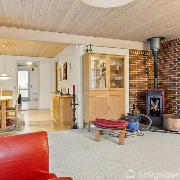 Rød lænestol i en stue med brændeovn, der står i hjørnet foran en murstensvæg. Ved siden af er et vitrineskab. I baggrunden ses et spisebord og stole.