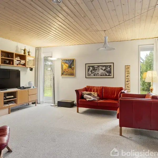 Rødt sofasæt står i en lys stue med trægulv og loft. Tv på vægmonteret reol, vinduer til venstre, kunstværker på væggene og tæppe på gulvet.