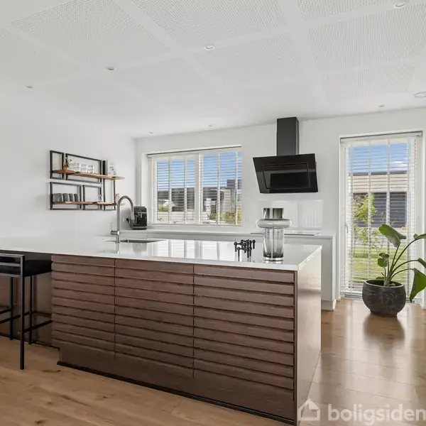 Et moderne køkken har en stor ø der fungerer som arbejdsstation med barstole. Sollys strømmer ind gennem store vinduer med persienner. En plante står ved siden af et skab.