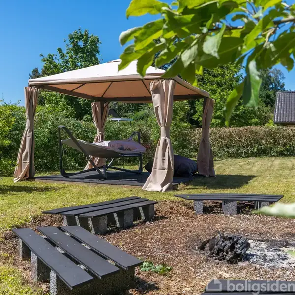 Pavillon med hvide gardiner står på en græsplæne. En hængesofa er placeret indenunder. I forgrunden ses bænke omkring et bålsted. Flot natur omgiver området.