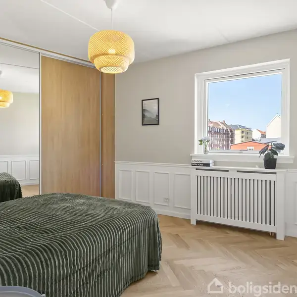Soveværelse med en grønstribet seng, der står stille. Store spejldøre klædeskab til venstre. Vindue viser udsigt til bygninger. Loftslampe hænger over sengen.