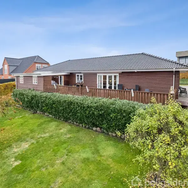 Et træhus med terrasse står på en velholdt græsplæne omgivet af buske. Den blå himmel og nabohuse ses i baggrunden.