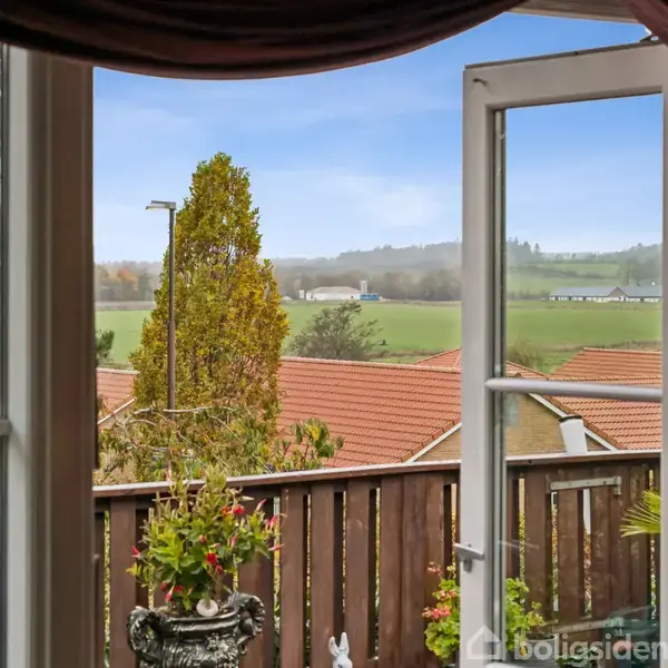 Et vindue åbner op til en udsigt over tage med røde tegl omgivet af marker og træer. En lygtepæl og en stor busk er synlig i forgrunden.