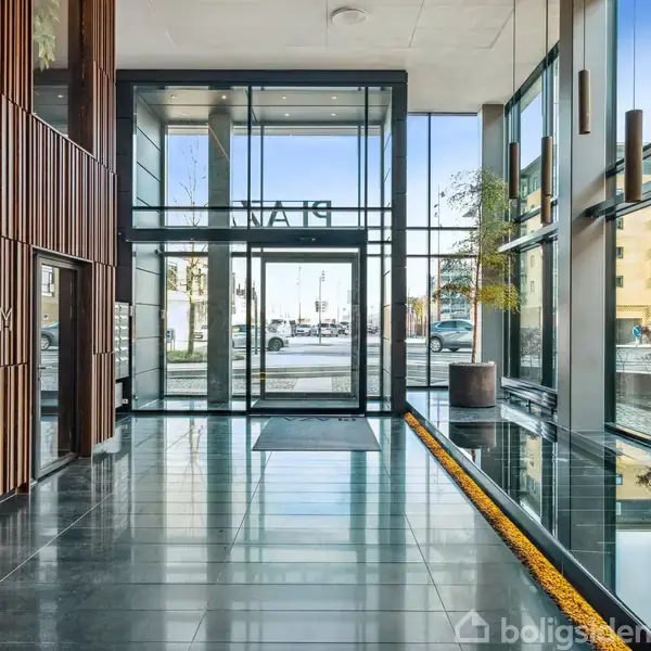 Lobby med glasdøre fører ud til gaden, omgivet af høje glasvægge. Træpanelvæg til venstre. Tekst: "CALUM PLAN". Bygninger og biler vises udenfor.